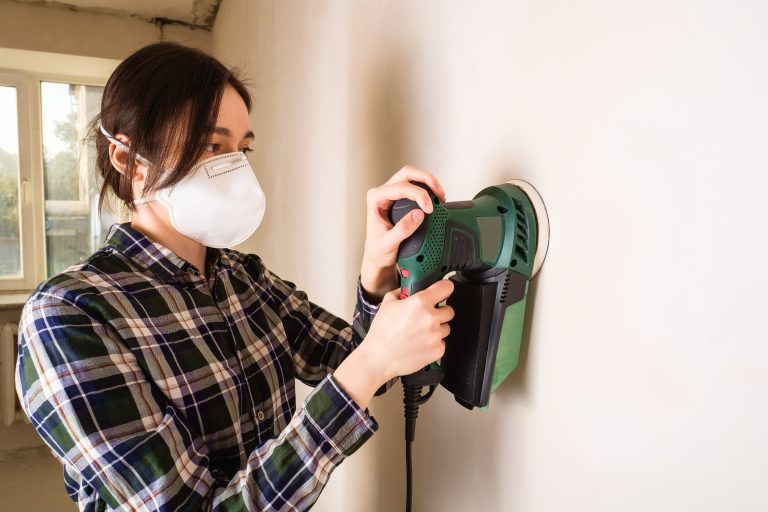 une femme portant un masque utilisant une ponceuse excentrique sur un mur placo